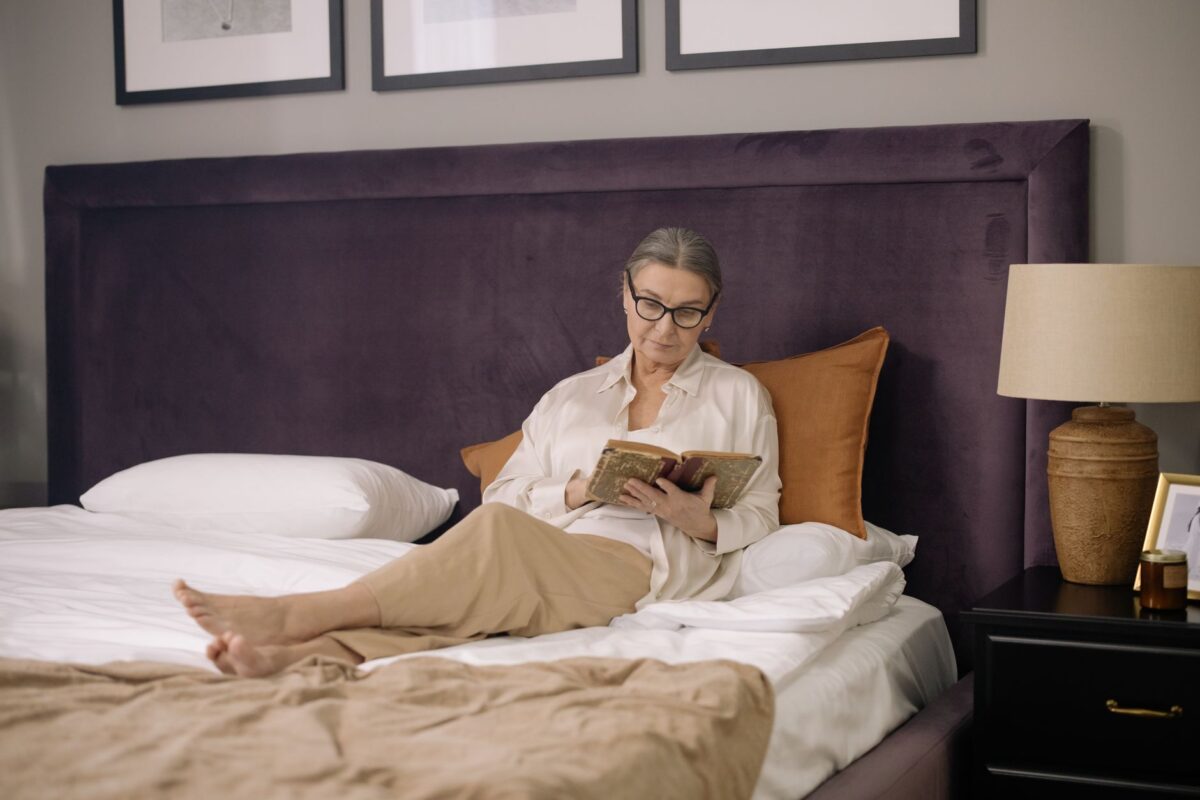 Woman reading a book on a bed beside a lit lamp in the evening