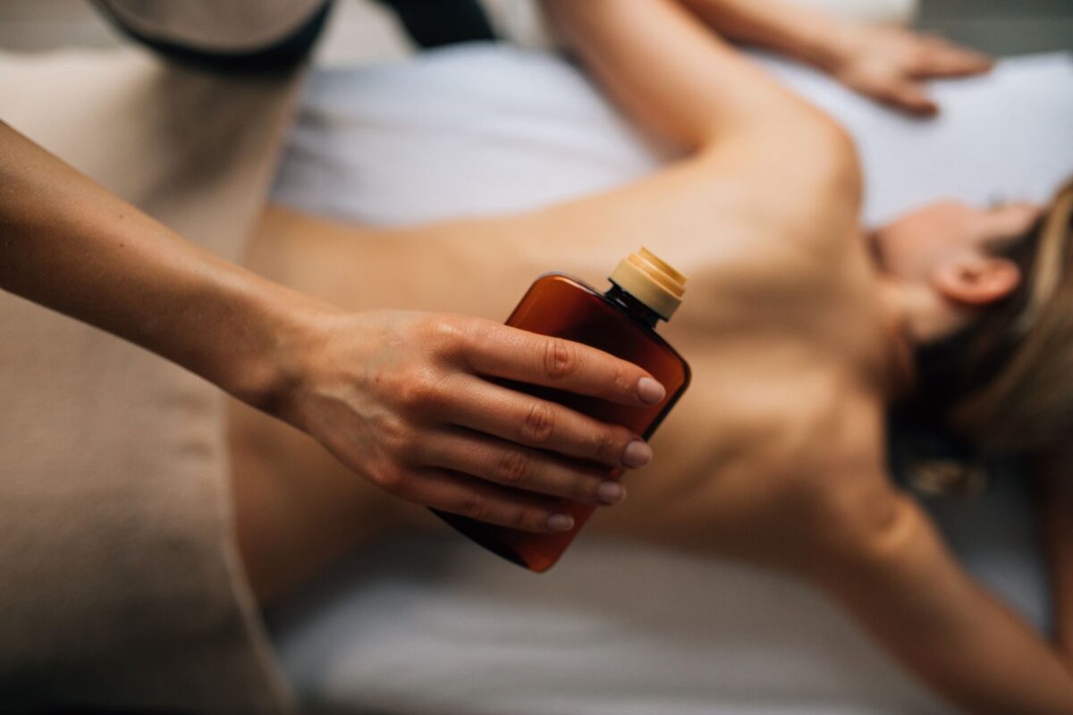 Therapist holding an amber bottle of massage oil over a client lying on a treatment table