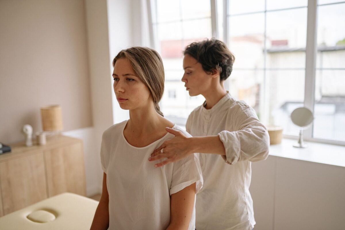 Therapist placing a light hand on a client's shoulder in a bright treatment room, both wearing linen and cotton