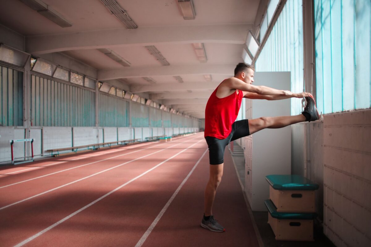 Athlete in a red top stretching his leg against a wall on an indoor running track
