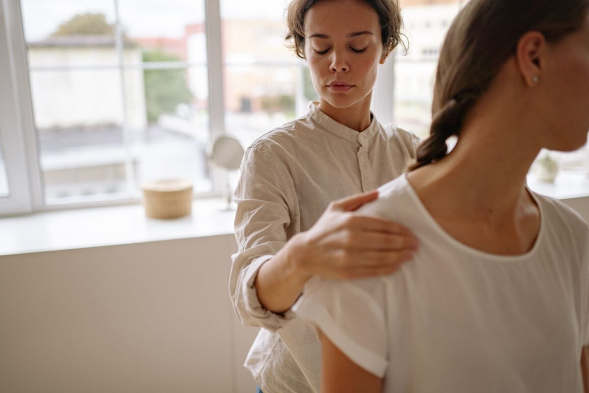 Practitioner with eyes closed resting a hand on a client's shoulder in a bright treatment room